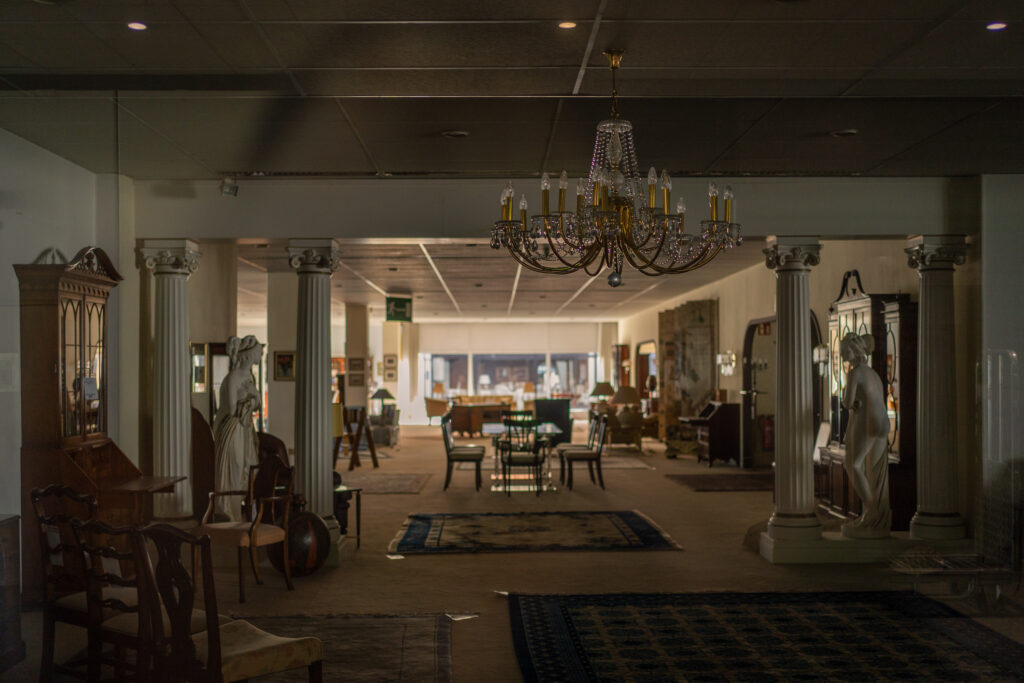 Blick in das Schaufenster des Möbelgeschäfts Hannelore Greve in Hamburg, City Nord. Es sind ältere Möbel im englischen Stil, im Vordergrund hängt ein goldener Kronleuchter. Das Bild ist in warmen Farben gehalten.